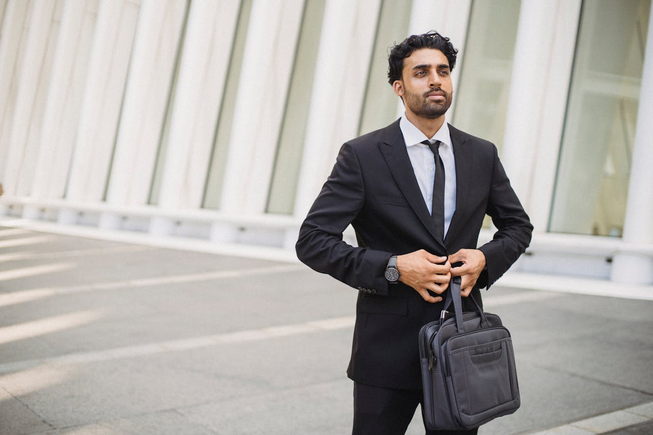 Home Business professional in a suit buttoning jacket while holding a briefcase outside a modern building.