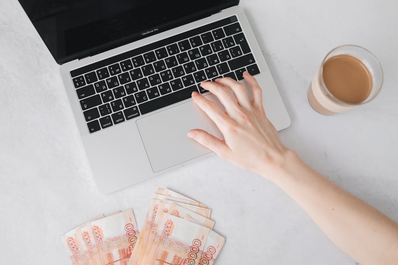 About Close-up of a hand on laptop keyboard with cash and coffee on table.