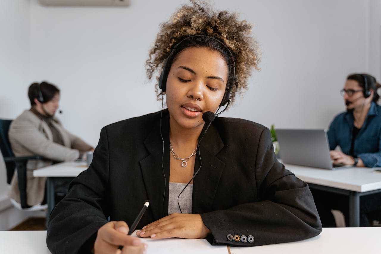 Services African American woman with headset working in an office with colleagues, using a pen for note-taking.